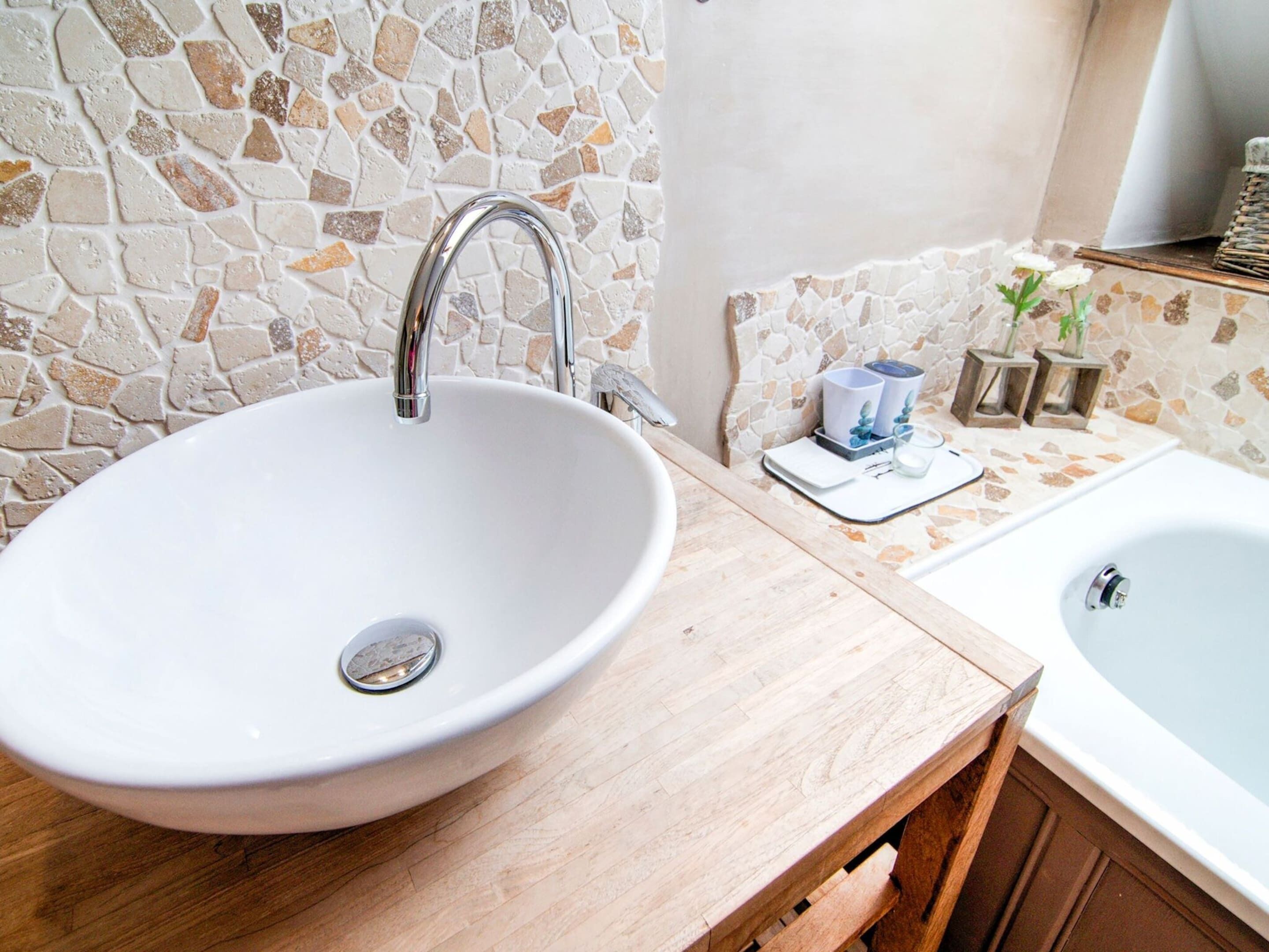 A bathroom with a white ceramic sink, a wooden vanity, and a stone mosaic backsplash.