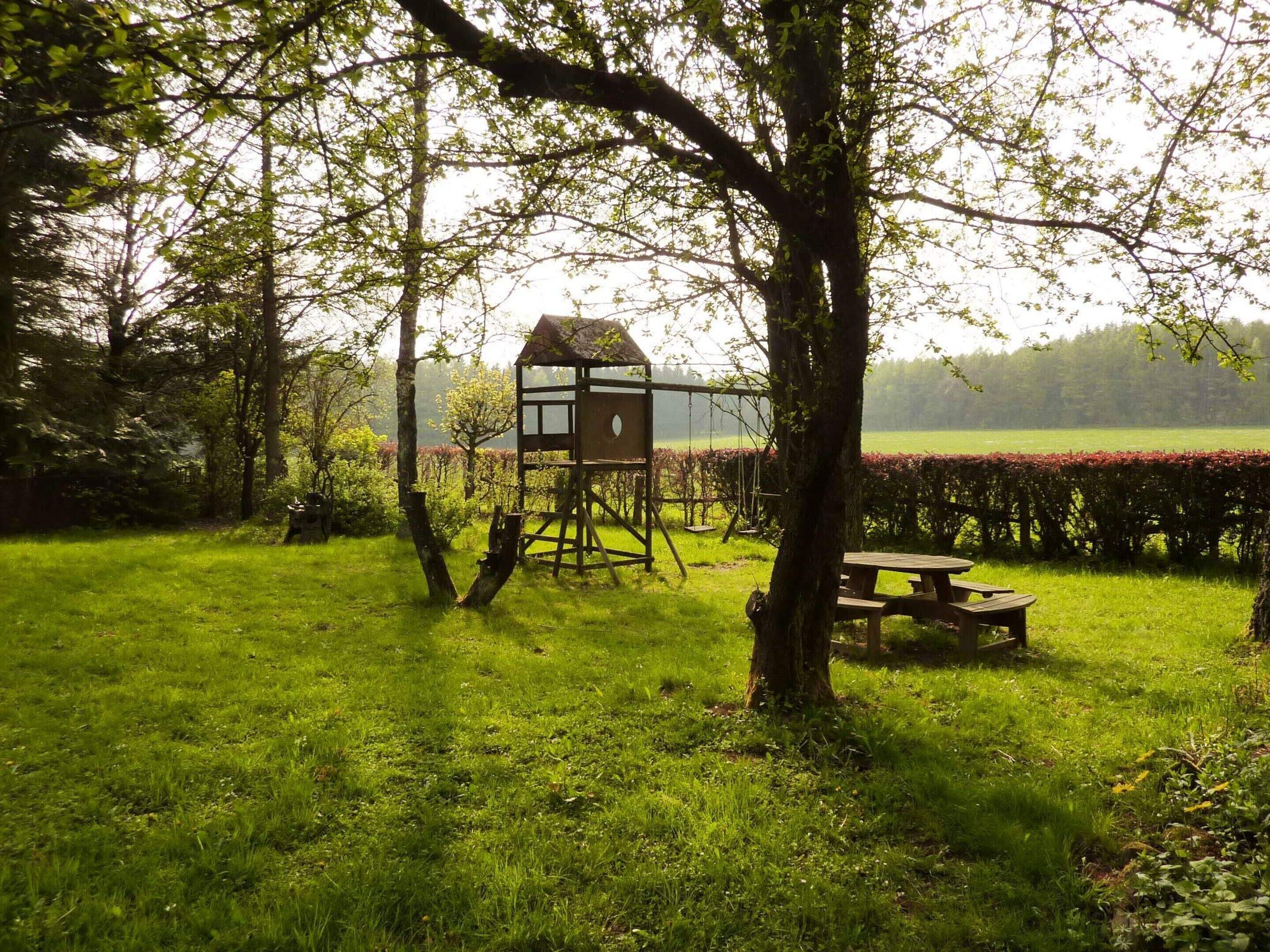 A rustic wooden playhouse with a swing set, surrounded by lush greenery and a picnic table.