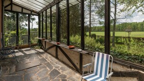 A covered patio with stone flooring, metal chairs, and a view of a green landscape through large windows. - Holiday Home in Waimes With Private Garden (Waimes)