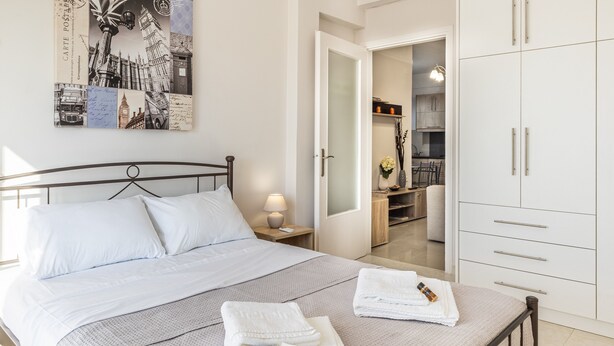 A neatly made bed with white linens and a folded towel on the side. There is a nightstand with a lamp and a framed picture on the wall. A white wardrobe is visible in the background. - Comfy Central Square Gem with Terrace (Kalamata)