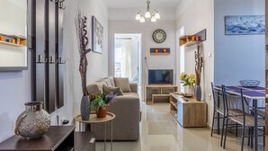 A modern living room with a sofa, TV, dining table, and chairs. There is a clock on the wall and a vase with flowers on a side table. - Comfy Central Square Gem with Terrace (Kalamata)