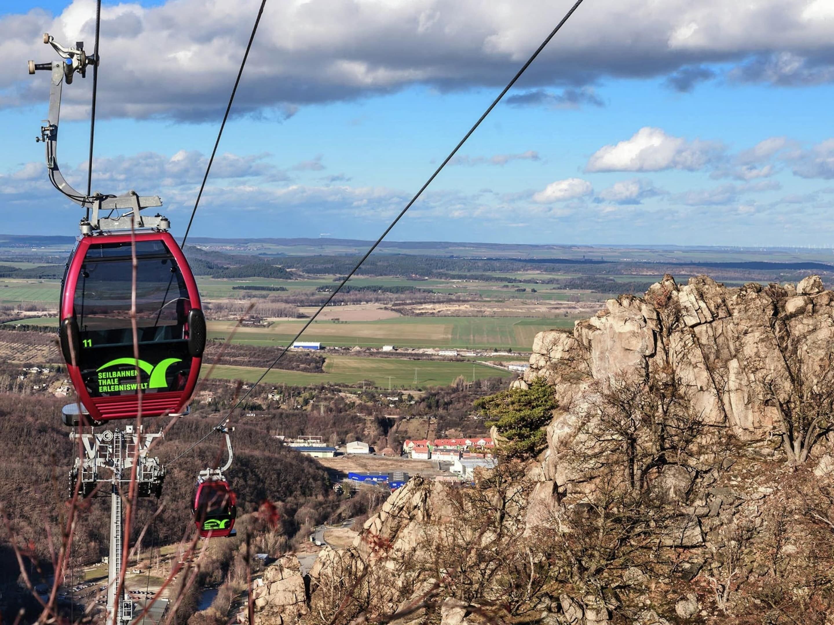 A cable car with a green logo, suspended above a rocky landscape with buildings in the distance.