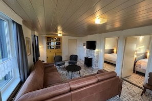 A room with a brown leather sofa, two black chairs, a small table, a TV, and a bed with a patterned rug.