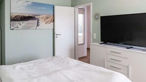 A bedroom with a bed, a TV, a door, and a painting of a beach scene. - Ferienhaus in Hasselfelde (Oberharz am Brocken)