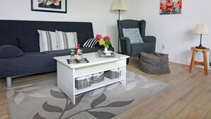 A living room with a dark blue sofa, a white coffee table, a grey armchair, and a small wooden stool. There is a framed picture on the wall and a lamp on a side table. - Cozy Apartment in Neubukow near Water Sports (Neubukow)