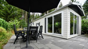 En modern uteplats med ett bord och stolar, ett svart parasoll och en vit byggnad med stora fönster.