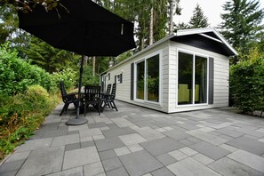 A patio with a table and chairs, a black umbrella, and a small building with large windows. - Cosy Chalet w/ Garden Patio (Vorden)