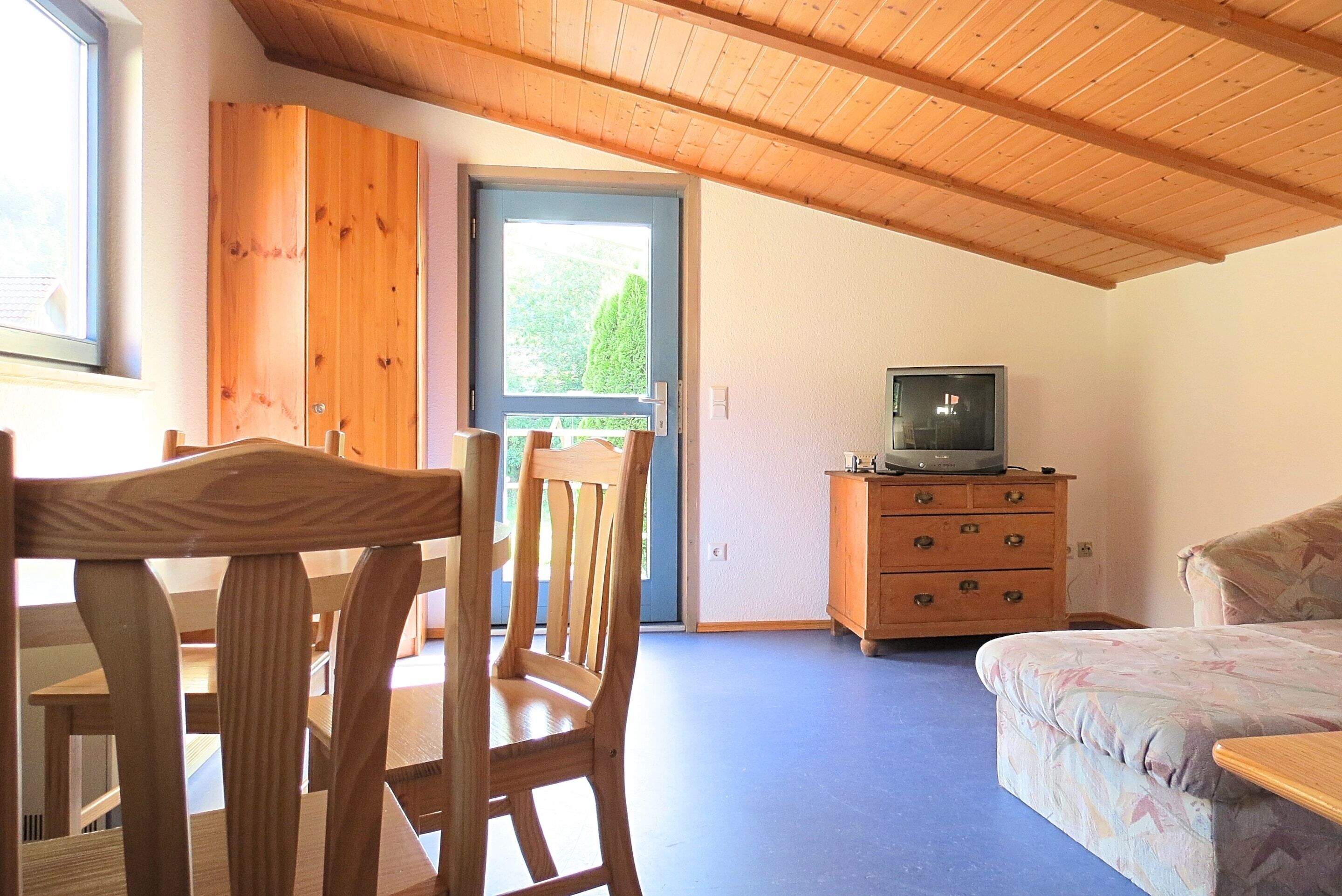 Quarto com teto de madeira, televisão sobre um móvel de madeira, uma cama e janela com vista para o verde.