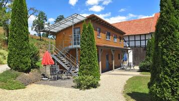 Uma casa de madeira de dois andares com telhado de telhas vermelhas, área de estar coberta ao ar livre e entrada com calçamento de pedras.