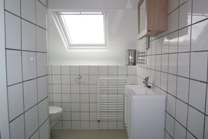 Une petite salle de bain carrelée, dotée d’un puits de lumière, de toilettes, d’un lavabo et d’un radiateur.