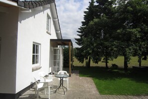 Um pátio com mesa e cadeiras, área coberta e gramado com árvores.