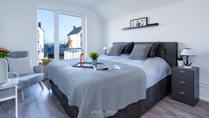 A modern bedroom with a large bed, a bedside table, a chair, and a view of the outside. - Ostseeskipper - Whirlpool, Shower, Fireplace, Infrared Sauna, 1st Floor, Grill (Olpenitz)