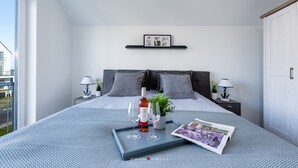 A modern bedroom with a large bed, two bedside tables with lamps, a small potted plant, a tray with wine glasses, and a magazine. - Ostseeskipper - Whirlpool, Shower, Fireplace, Infrared Sauna, 1st Floor, Grill (Olpenitz)