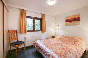 A bedroom with a bed, a chair, a window with curtains, a radiator, a wall-mounted lamp, and a painting.