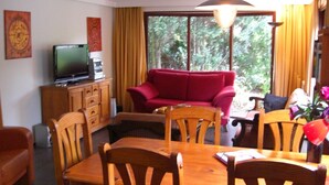 Un salon avec un canapé rouge, une table à manger en bois et des chaises, un téléviseur posé sur un meuble et une grande fenêtre avec des rideaux.
