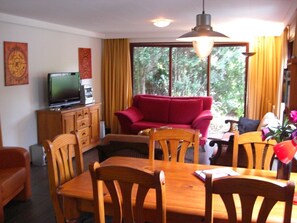 A living room with a red sofa, wooden dining table, and chairs, a television on a cabinet, and a large window with curtains.