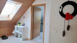 A room with a sloped ceiling, a skylight, a white shelf with a potted plant, a door leading to another room, and a wall decoration with a circular frame and hanging elements. - Attic Apartment, Herrischried (Herrischried)