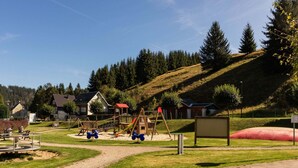 Un terrain de jeux avec des balançoires et des toboggans, entouré de maisons et d’un paysage vallonné.