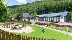 Une aire de jeux avec des balançoires et des toboggans, un bâtiment doté de panneaux solaires et, au loin, une colline boisée.