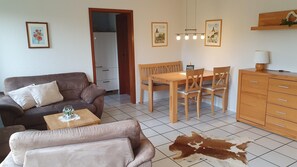 A living room with a brown sofa, a small table with a glass vase, a dining area with a wooden table and chairs, and a kitchen area with wooden cabinets and a hanging light fixture.