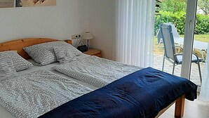 Un dormitorio con cama, mesita de noche, lámpara y vista a un jardín a través de un ventanal.