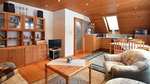 A cozy, well-lit room with wooden ceilings and walls, a television, a dining area with a table and chairs, and a kitchen area with a microwave and toaster. - Holiday Home Beerheide Near Ski Slopes (Auerbach)
