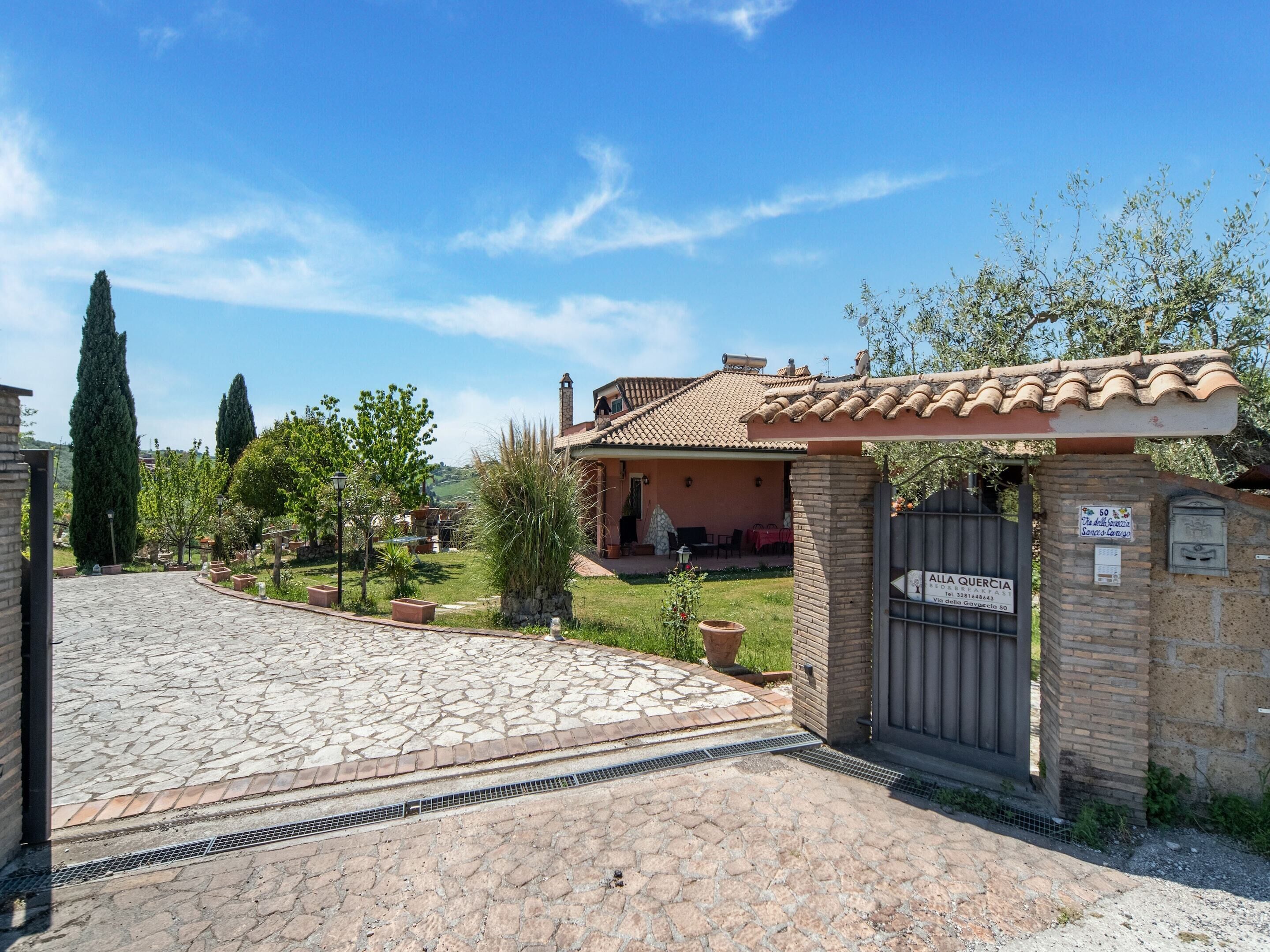 A house with a paved driveway leading to a gated entrance, surrounded by greenery and a clear sky.