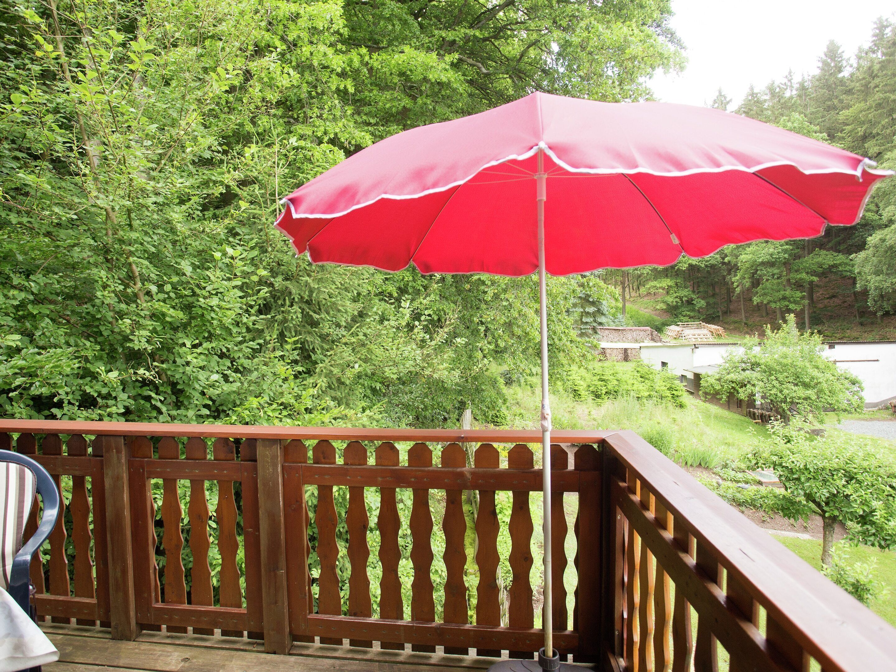 A wooden deck with a red umbrella, surrounded by dense green foliage and a white building in the background.