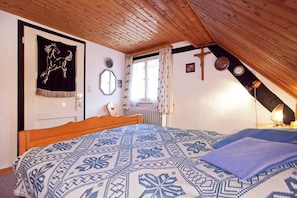 Ein Schlafzimmer mit schrägem Dach, einem Bett mit gemusterten Textilien, einem Holz-Kopfteil, einem Fenster mit Vorhängen und einer Wanduhr.