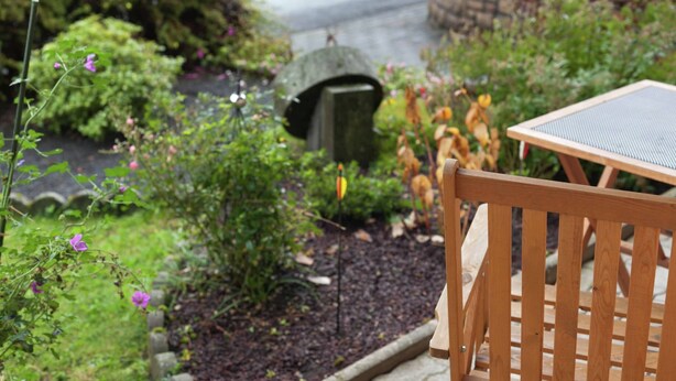 A wooden outdoor seating area with a table and chair, surrounded by a garden with various plants and a small pond. - Einladende Ferienwohnung in Muellenborn Eifel (Gerolstein)