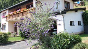 A two-story building with a balcony adorned with colorful flowers, surrounded by lush greenery and a parking area. - Einladende Ferienwohnung in Muellenborn Eifel (Gerolstein)