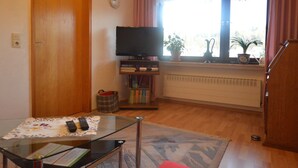 A living room with a television, books, and a window with curtains. - Einladende Ferienwohnung in Muellenborn Eifel (Gerolstein)