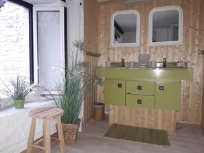 Interior em madeira com uma pia verde, dois espelhos, uma planta em vaso e um banquinho de madeira.
