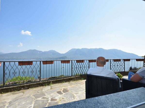 A terrace with a stone floor, metal railing, and a view of mountains and a lake.