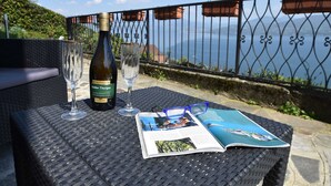 Ett bord med en vinflaska, glas och en tidning, placerat utomhus på ett bord av flätat material, med utsikt över havet.