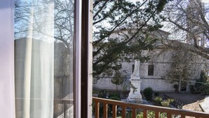 A view from a window with a wooden railing, overlooking a courtyard with a statue, a stone building, and trees. - Secluded Mountain Refuge - Theonimfi Paradise Gem (Gortynia)