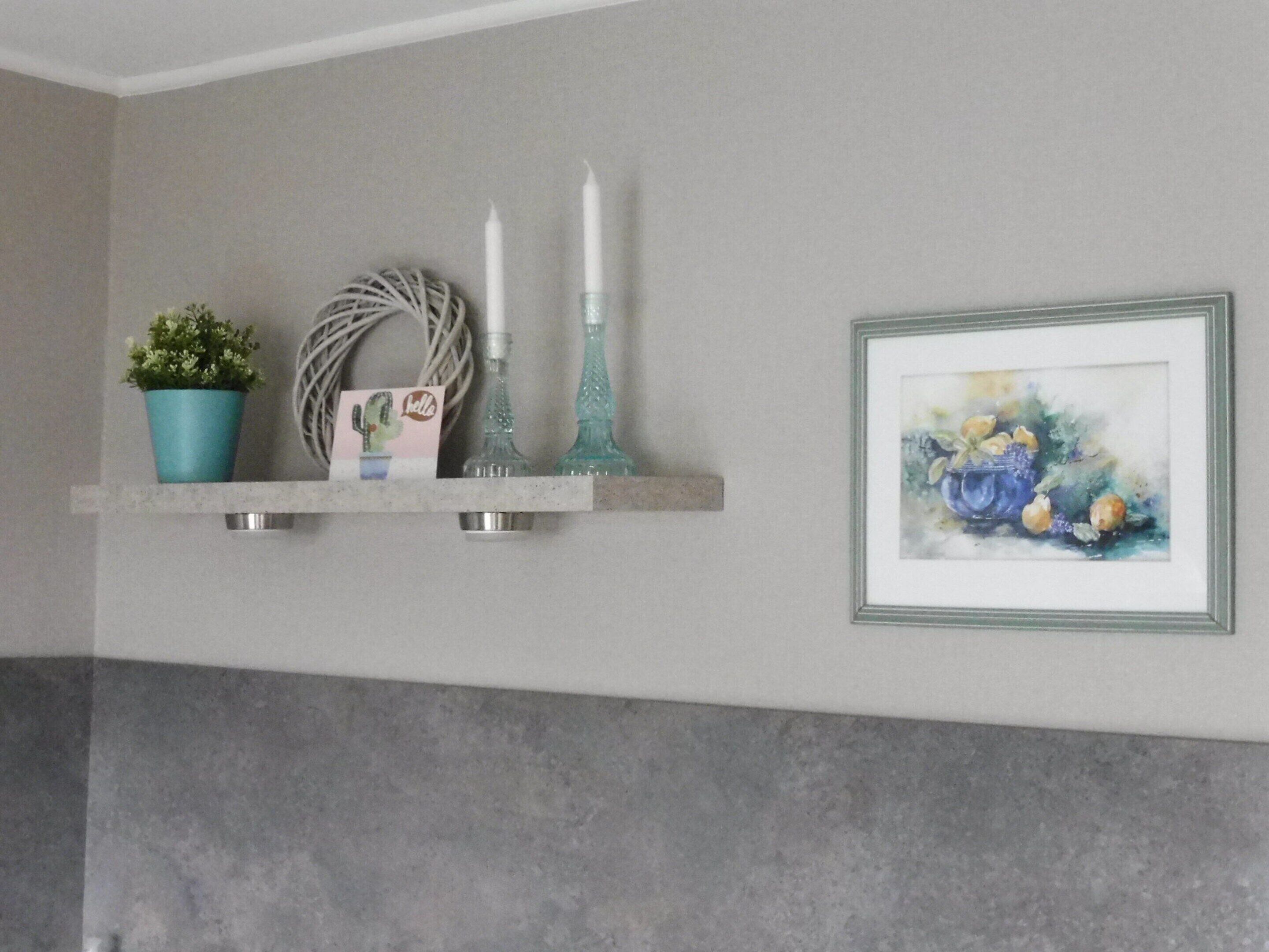 A bathroom with a grey wall, a white ceiling, and a grey floor. There is a shelf with a potted plant, a candle, and a framed picture.