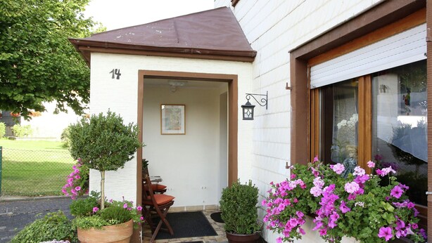 A building with a white exterior, a brown roof, and a door with a small porch area. There are potted plants and flowers outside. - Spacious Apartment in Pracht near Forest (Pracht)