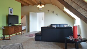 A living room with a wooden ceiling, a black sofa, a television on a wooden cabinet, and a red vase on a table. - Farmstay Comfort in Juchen (Juechen)