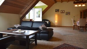 A cozy living room with a dark leather sofa, a wooden dining table, and a high ceiling with exposed beams. - Farmstay Comfort in Juchen (Juechen)