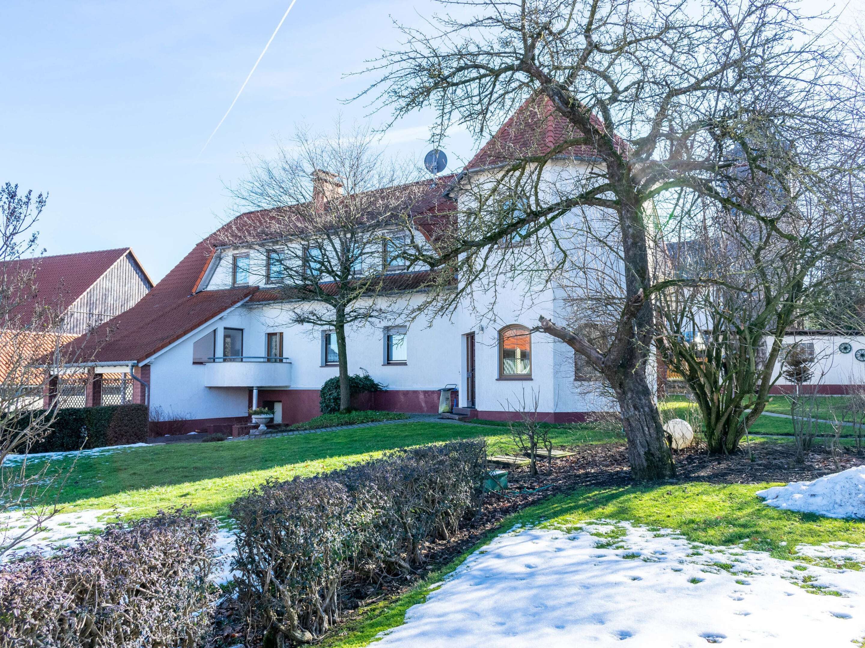 A two-story house with a red roof, white walls, and a garden with snow on the ground.