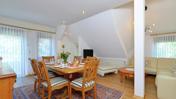 A dining area with a wooden table, chairs, and a patterned rug. There is a sofa and a small table in the background. - Quaint Apartment in Eimelrod near Lake & Water Sports (Willingen)