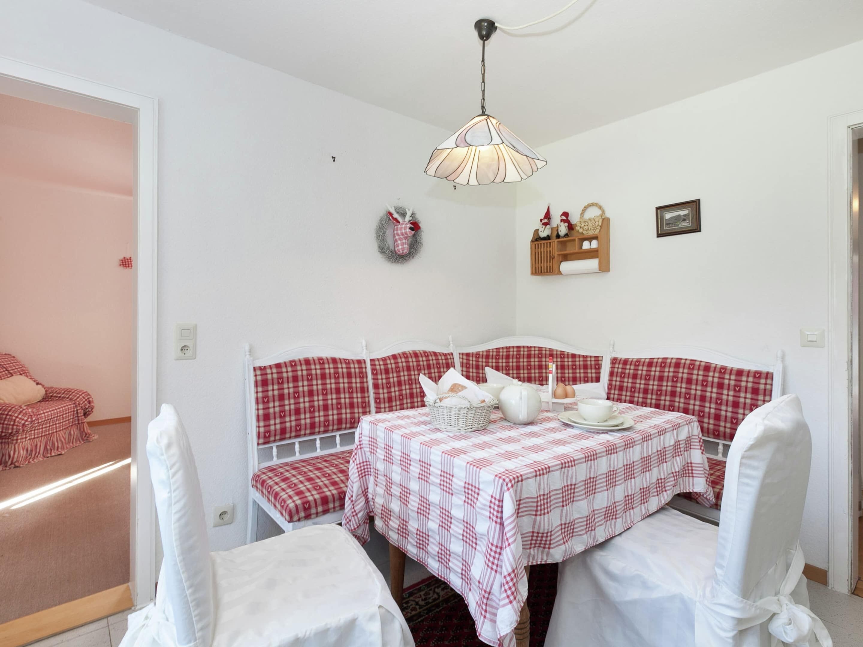 A dining area with a checkered tablecloth, white chairs, a white bench, a chandelier, and a wall-mounted shelf with decorative items.