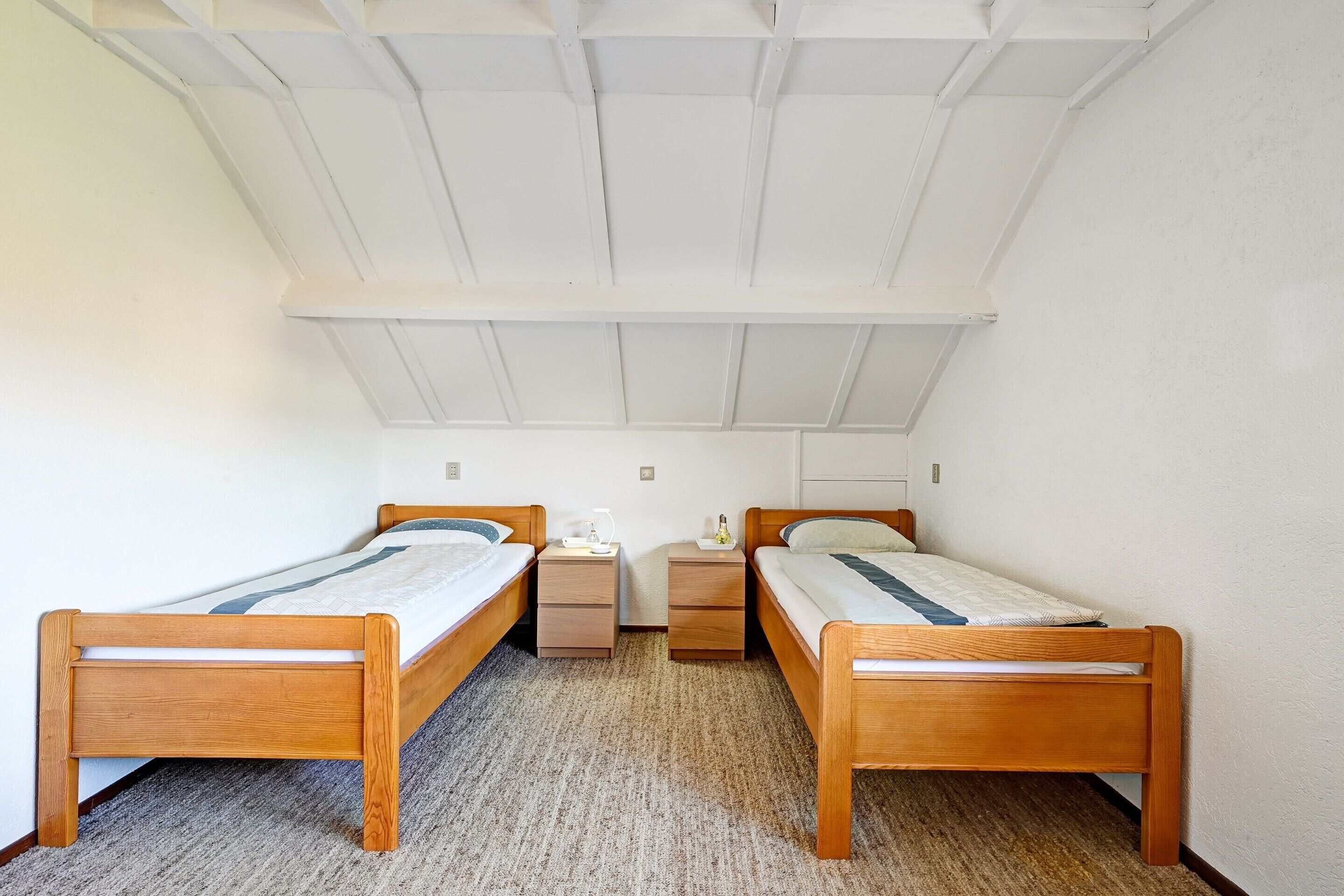 A small, clean room with two wooden beds, a small bedside table, and a ceiling with wooden beams.