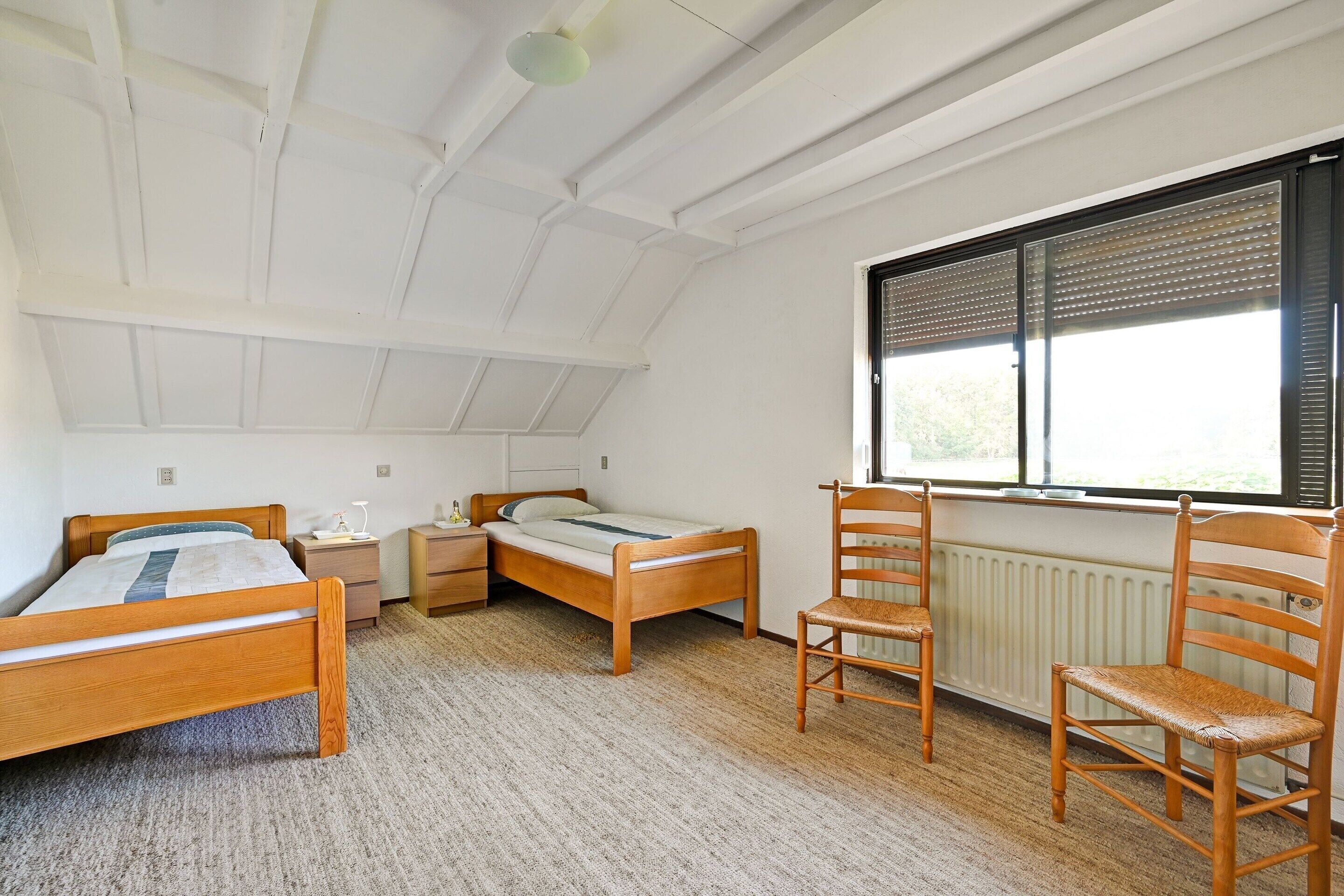 Una habitación con techo inclinado, dos camas de madera, una silla de madera, una mesita de noche y una ventana con persianas.
