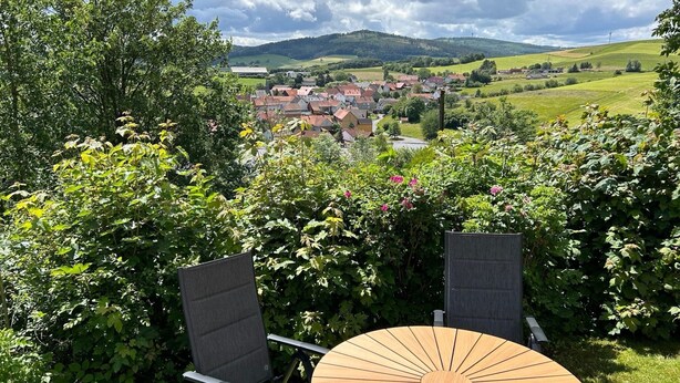 A patio with two chairs and a round table, overlooking a village with houses and hills.