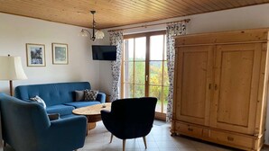 A living room with a blue sofa, a black armchair, a wooden coffee table, a TV mounted on the wall, and a large wooden wardrobe.
