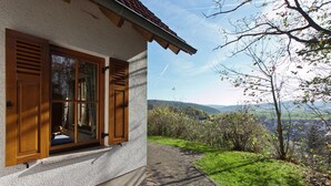 A house with a large window and wooden shutters, a paved pathway, and a view of a green landscape with trees and hills.