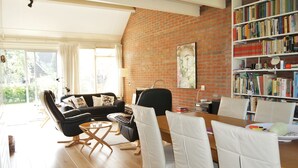 A living room with a black leather sofa, white chairs, a wooden dining table, and a brick wall with a bookshelf.