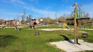 A playground with various play structures, including swings and climbing frames, surrounded by a grassy area with houses in the background.